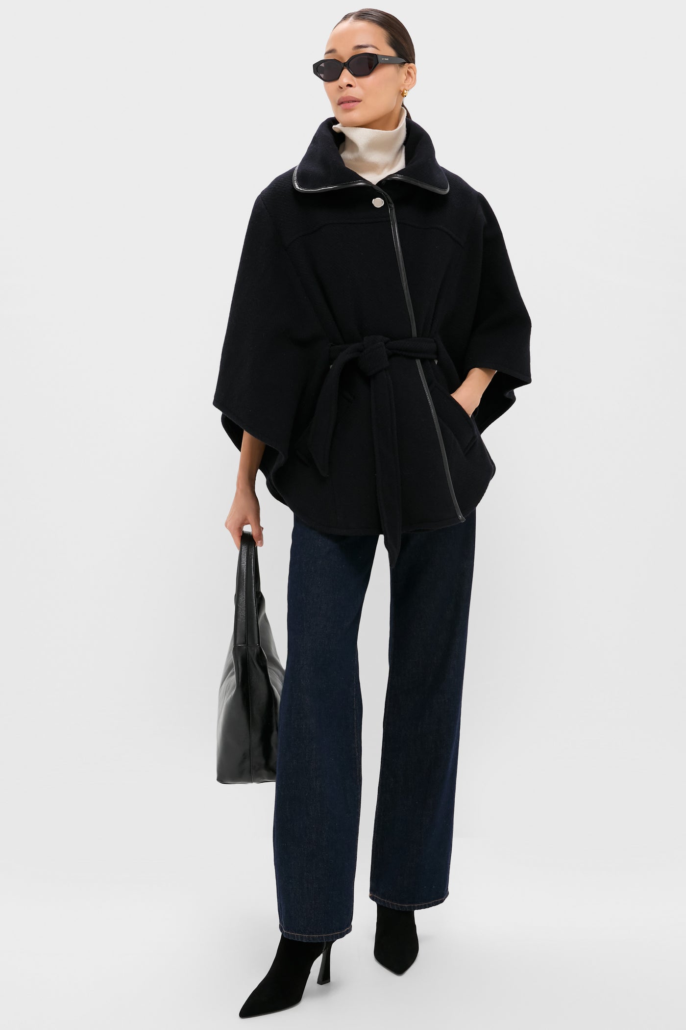 Wearing a ba&sh black belted wool cape coat, dark jeans, black heeled boots, sunglasses, and holding a black bag—an effortlessly chic nod to French fashion against a plain light background.