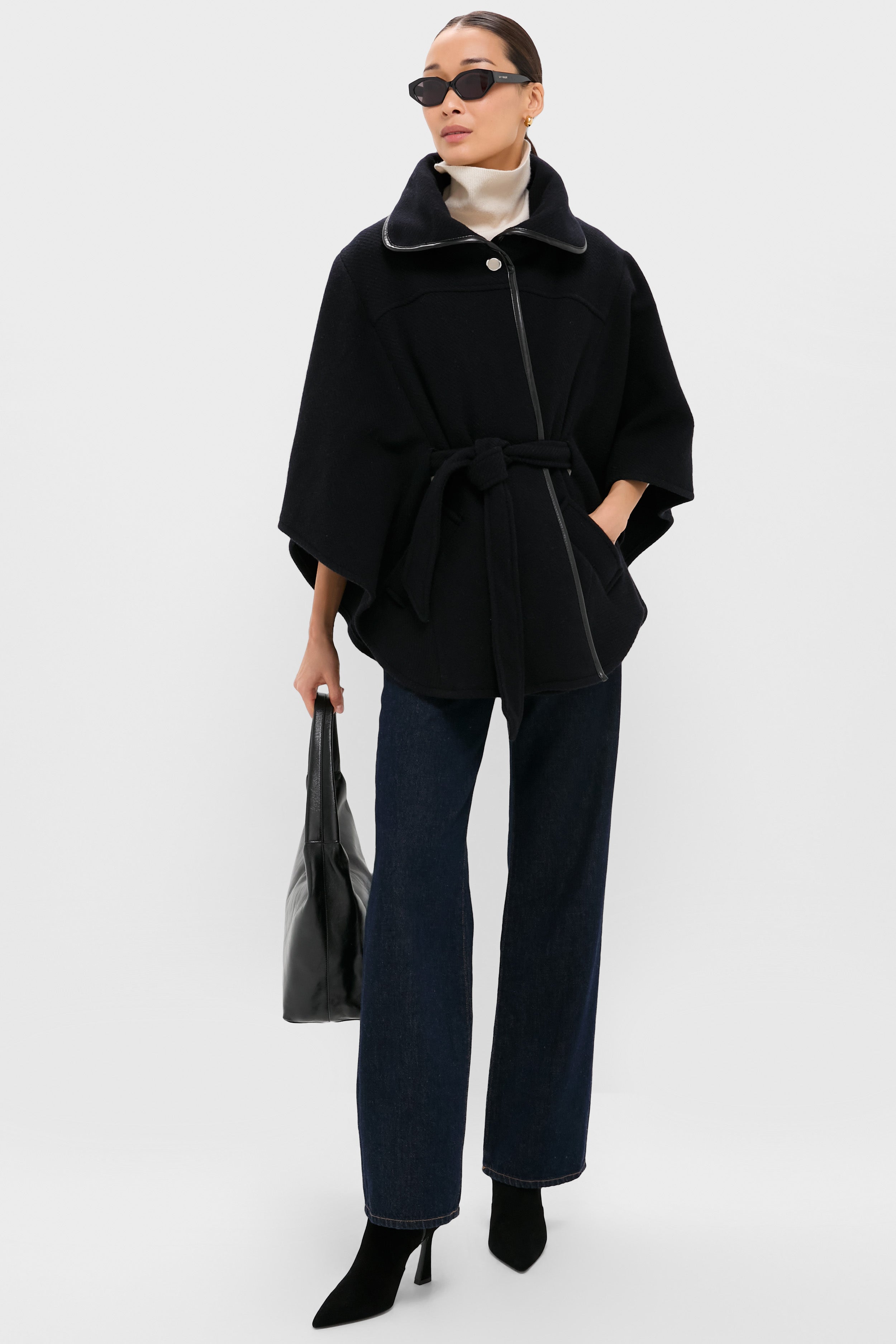 Wearing a ba&sh black belted wool cape coat, dark jeans, black heeled boots, sunglasses, and holding a black bag—an effortlessly chic nod to French fashion against a plain light background.