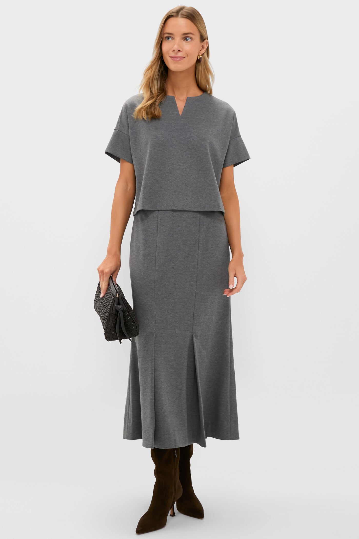 A woman in a Tuckernuck short-sleeved gray two-piece dress set with a notched neckline and cropped hem, holding a textured clutch and wearing dark brown knee-high boots.
