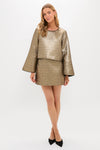 A woman models Tuckernuck’s gold jacquard mini skirt and matching metallic long-sleeve top with black trim, styled with black pointed-toe shoes against a white background—a stylish transitional wardrobe pick.