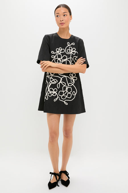 Woman in Onyx and Ivory Classic Katya Mini Dress with white floral embroidery, showcasing an effortless, high-end style; paired with fun, ribbon-tied velvet flats.