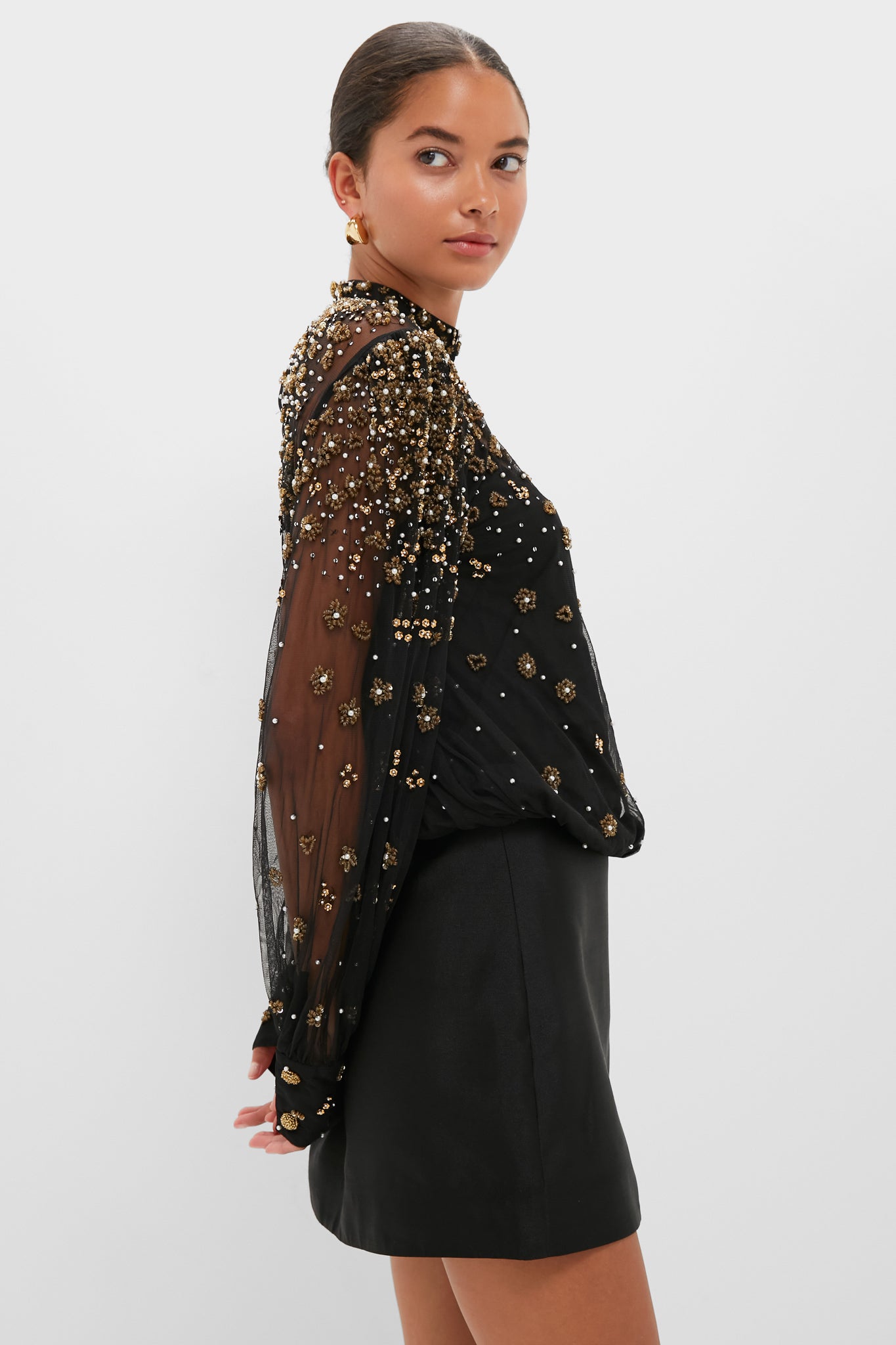 A woman wears a sheer Farm Rio top with gold embellishments and a black skirt, posing against a plain light background.