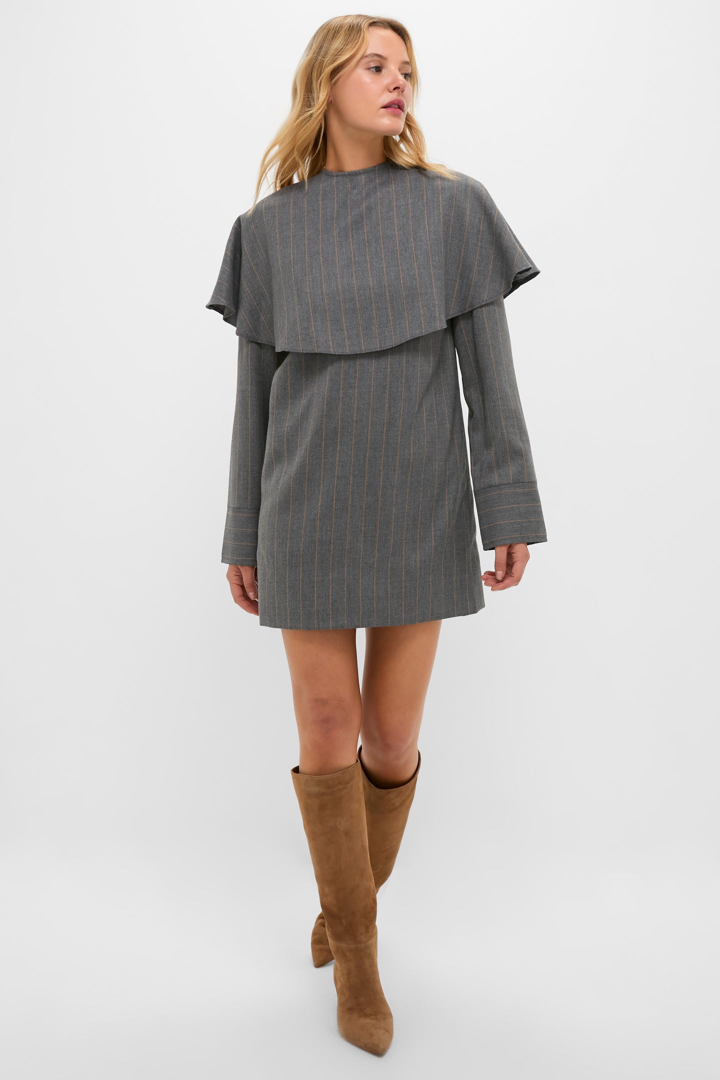 A woman stands against a white background wearing a GANNI mini dress with gray stripes and cape-style detailing, paired with tan knee-high suede boots.