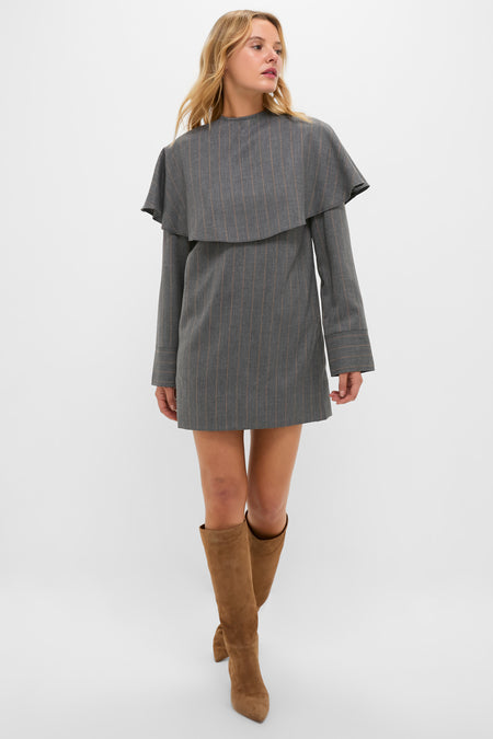A woman stands against a white background wearing a GANNI mini dress with gray stripes and cape-style detailing, paired with tan knee-high suede boots.