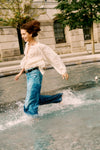 Woman kicking up water in a fountain in a white blouse and jeans. 