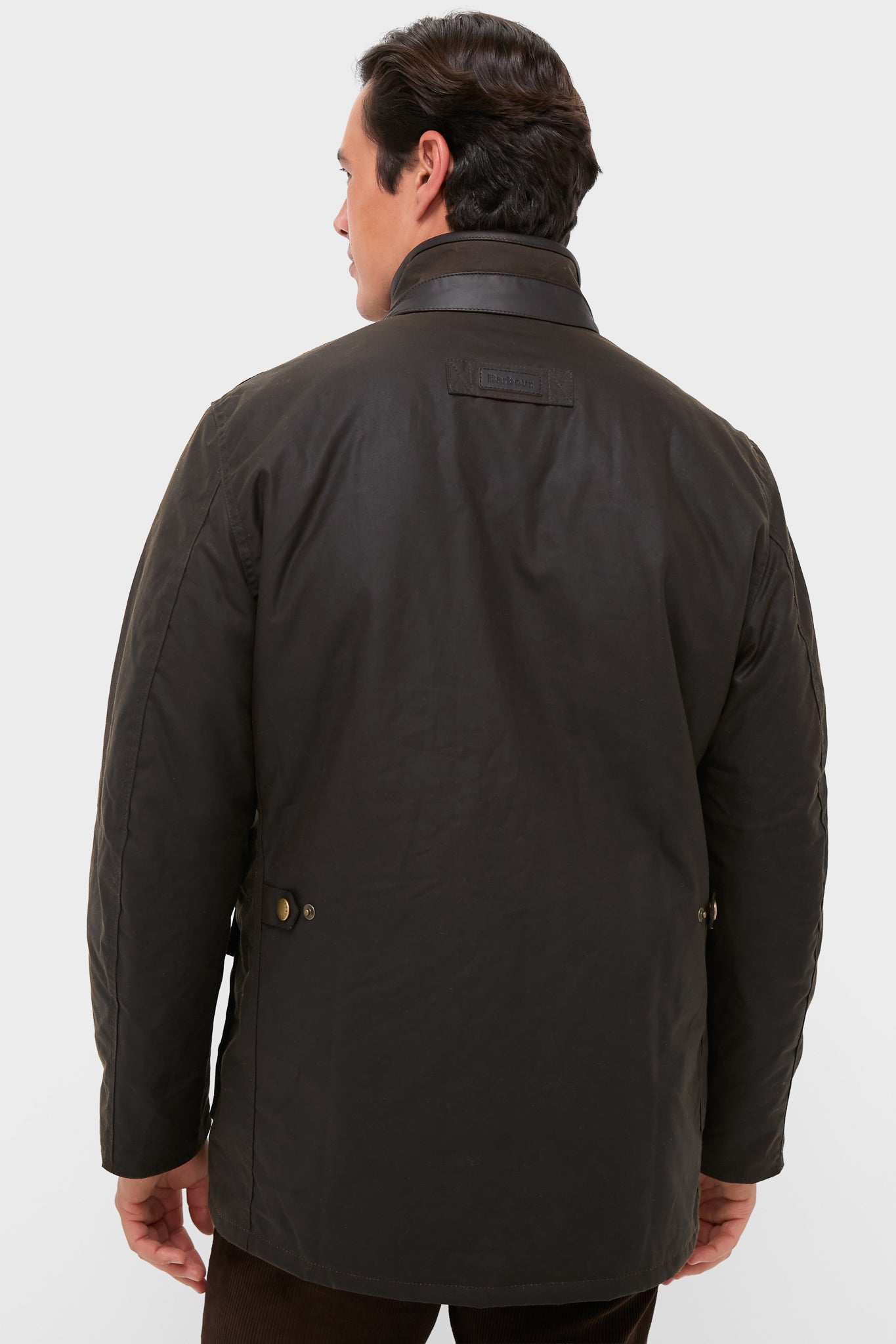 Back view of a man in a classic, high-end Olive Hereford Wax Jacket with leather trim, effortlessly showcasing Barbour’s timeless style and functional design against a light-gray backdrop.