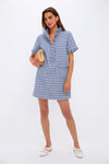 A woman in a Pomander Place Blue Tweed Ophelia Dress with fringe details holds a gold clutch and stands in white sneakers against a plain white background.