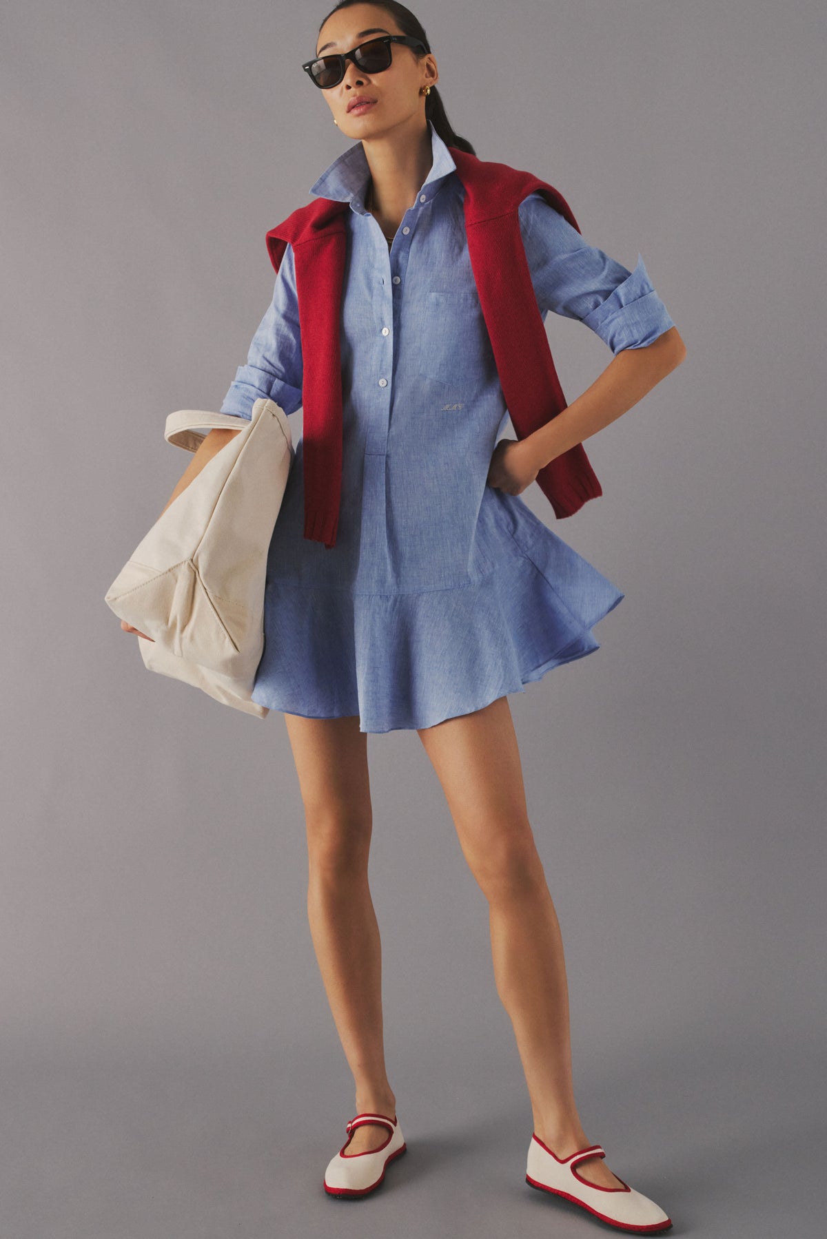 Woman standing in a studio in a chambray mini dress with a small white monogram on the waist. 
