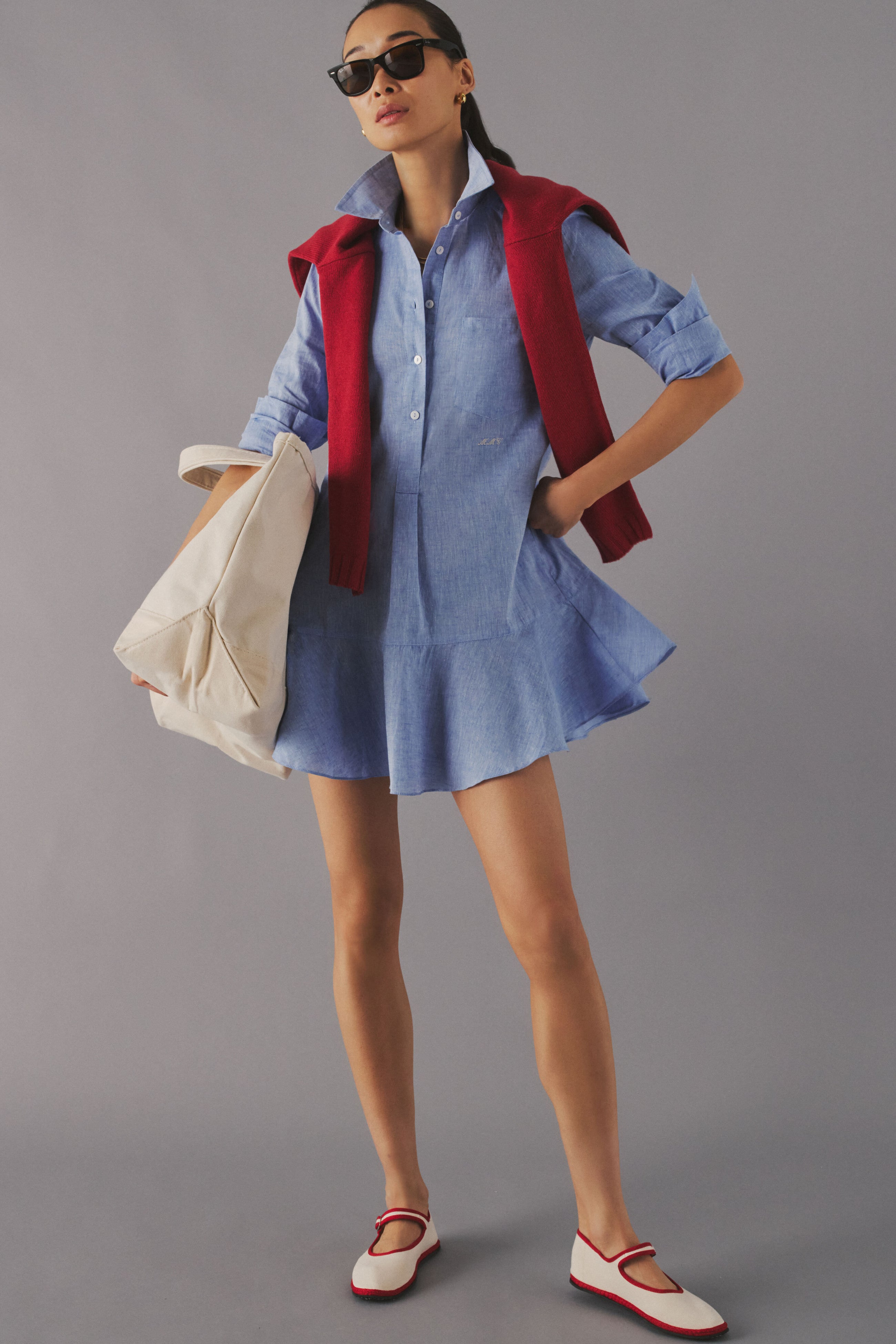 Woman standing in a studio in a chambray mini dress with a small white monogram on the waist. 