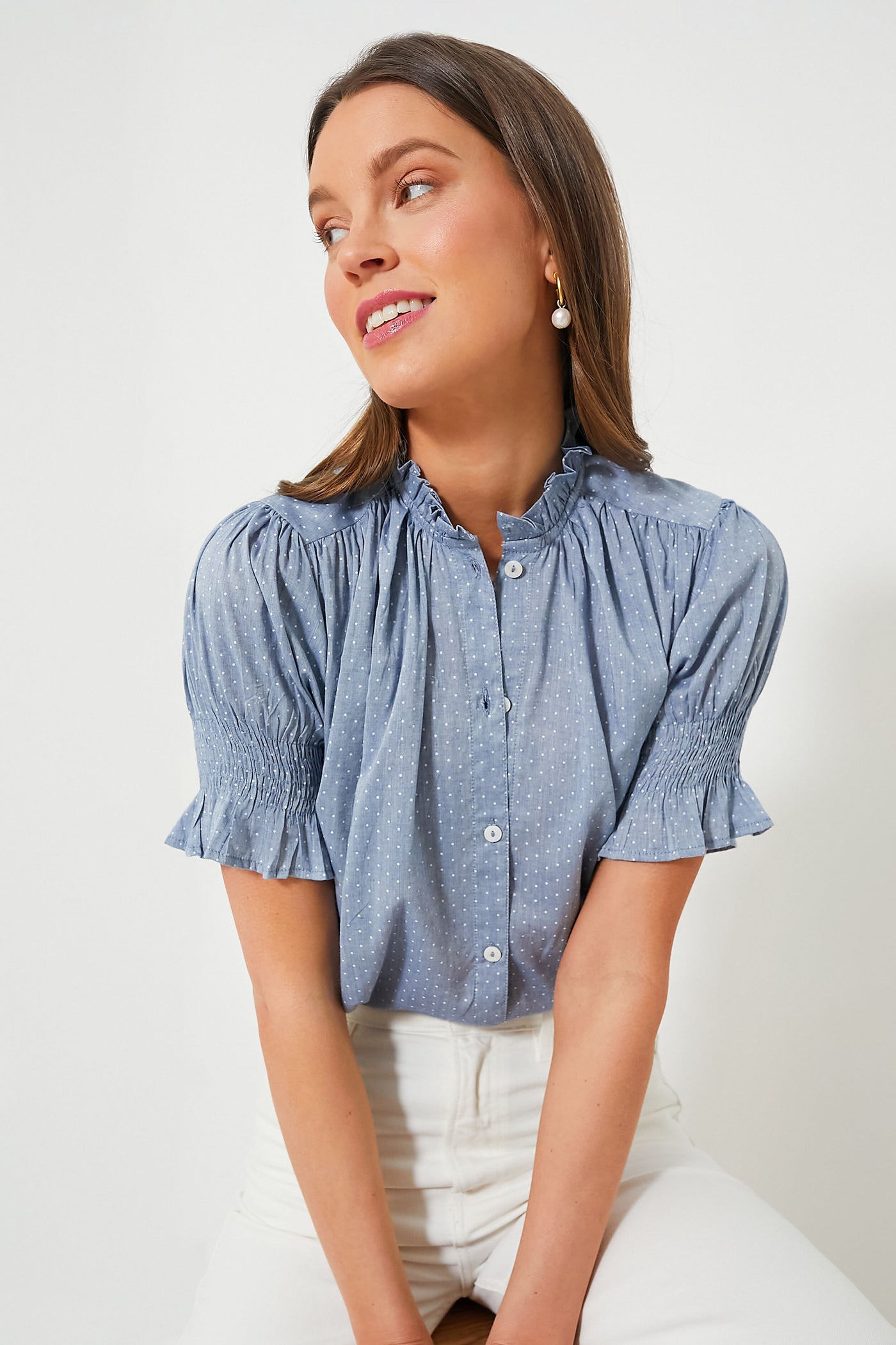 A woman seated and looking to her left wears the Mille Chambray Polka Dot Marnie Top featuring short puff sleeves and a ruffle neckline, paired with white pants.