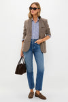 A woman wears Tuckernuck sunglasses, a chestnut herringbone blazer over a striped shirt, blue jeans, loafers, and carries a brown handbag while standing against a plain background.