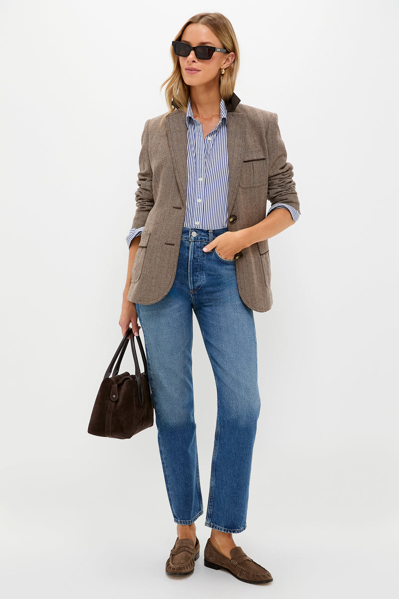 A woman wears Tuckernuck sunglasses, a chestnut herringbone blazer over a striped shirt, blue jeans, loafers, and carries a brown handbag while standing against a plain background.