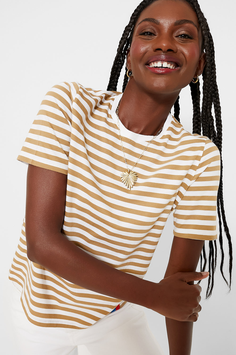 A woman wearing a KULE beige and white striped short-sleeve tee, 100% cotton white pants, gold hoop earrings, and a necklace smiles pleasantly.