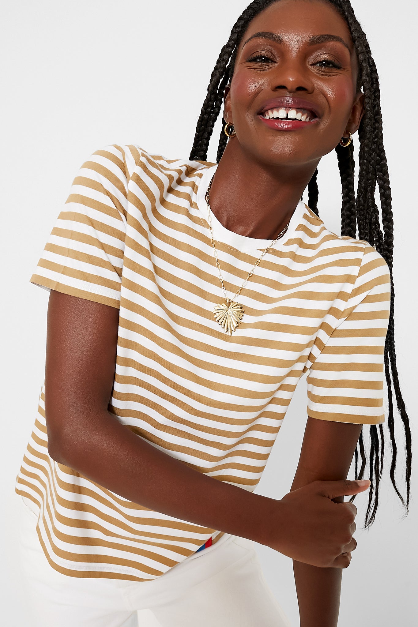 A woman wearing a KULE beige and white striped short-sleeve tee, 100% cotton white pants, gold hoop earrings, and a necklace smiles pleasantly.