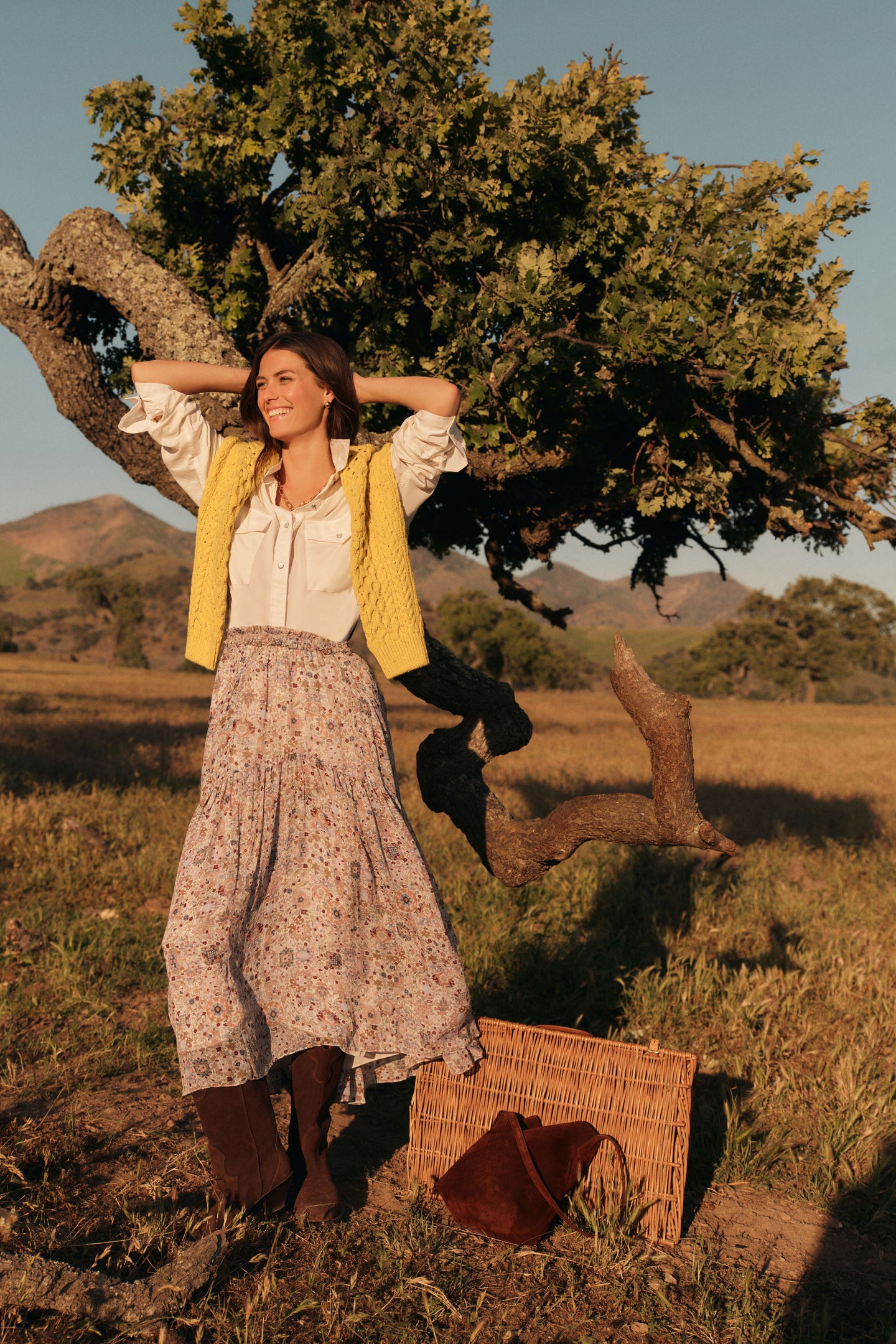 Woman standing outside in a long sleeved white button down shirt with the sleeves rolled up, a yellow cable knit sweater around her shoulders, a floral patterned maxi skirt, and chocolate brown boots. 