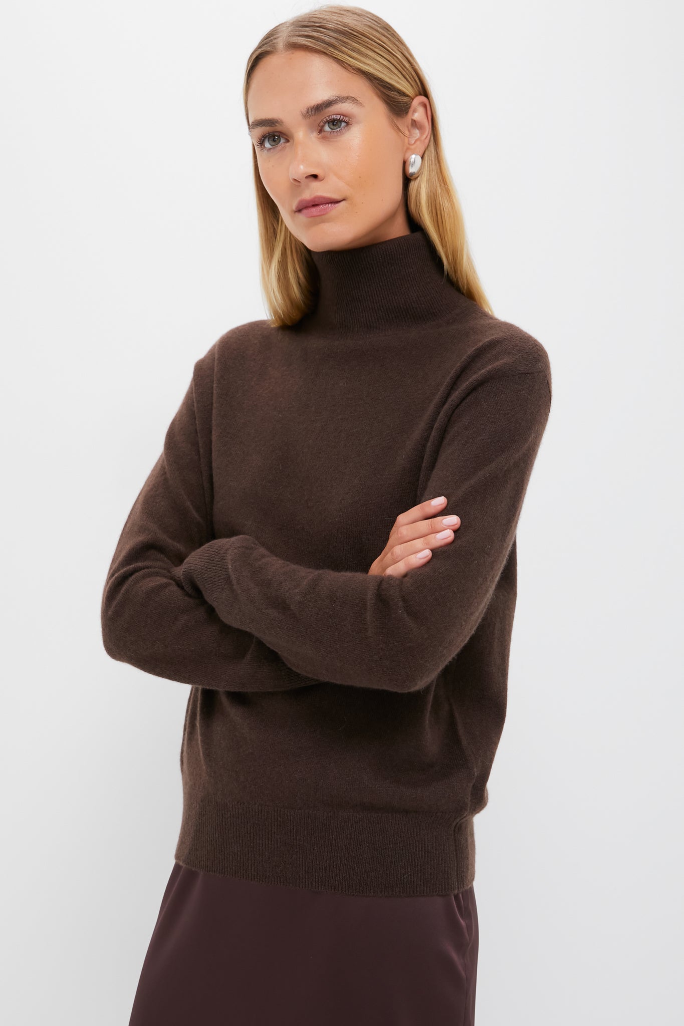 A woman with straight blonde hair wears a cozy Tuckernuck cashmere turtleneck sweater and pearl earrings, standing with her arms crossed against a plain white background.