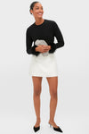 A woman stands wearing a black long-sleeve top with feather cuffs, a white mini skirt, black heels, and holds a sparkly clutch. She accessorizes with elegant Silver Klara Earrings by Lie Studio.