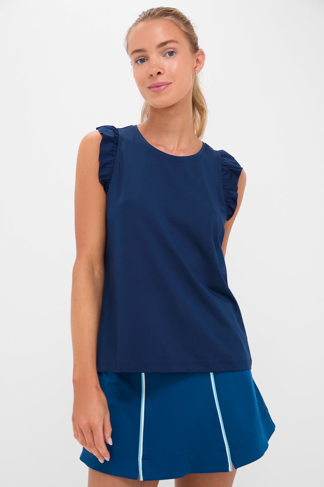 A woman models a sporty athleisure look in Tnuck Sport’s navy sleeveless tennis top with ruffled shoulders and a blue skirt featuring white piping.