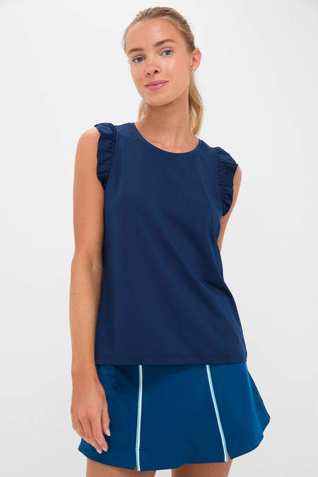 A woman models a sporty athleisure look in Tnuck Sport’s navy sleeveless tennis top with ruffled shoulders and a blue skirt featuring white piping.