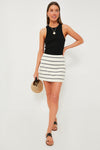 A woman stands against a white background wearing a Tuckernuck black sleeveless top, Tuckernuck Navy Stripe Mini Skirt, black sandals, and holding a round straw handbag.
