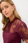 Young woman models the Oxblood Lace Agatha Mini Dress featuring a sheer lace yoke and sleeves, paired with chunky gold-tone earrings for a classic, effortless look.