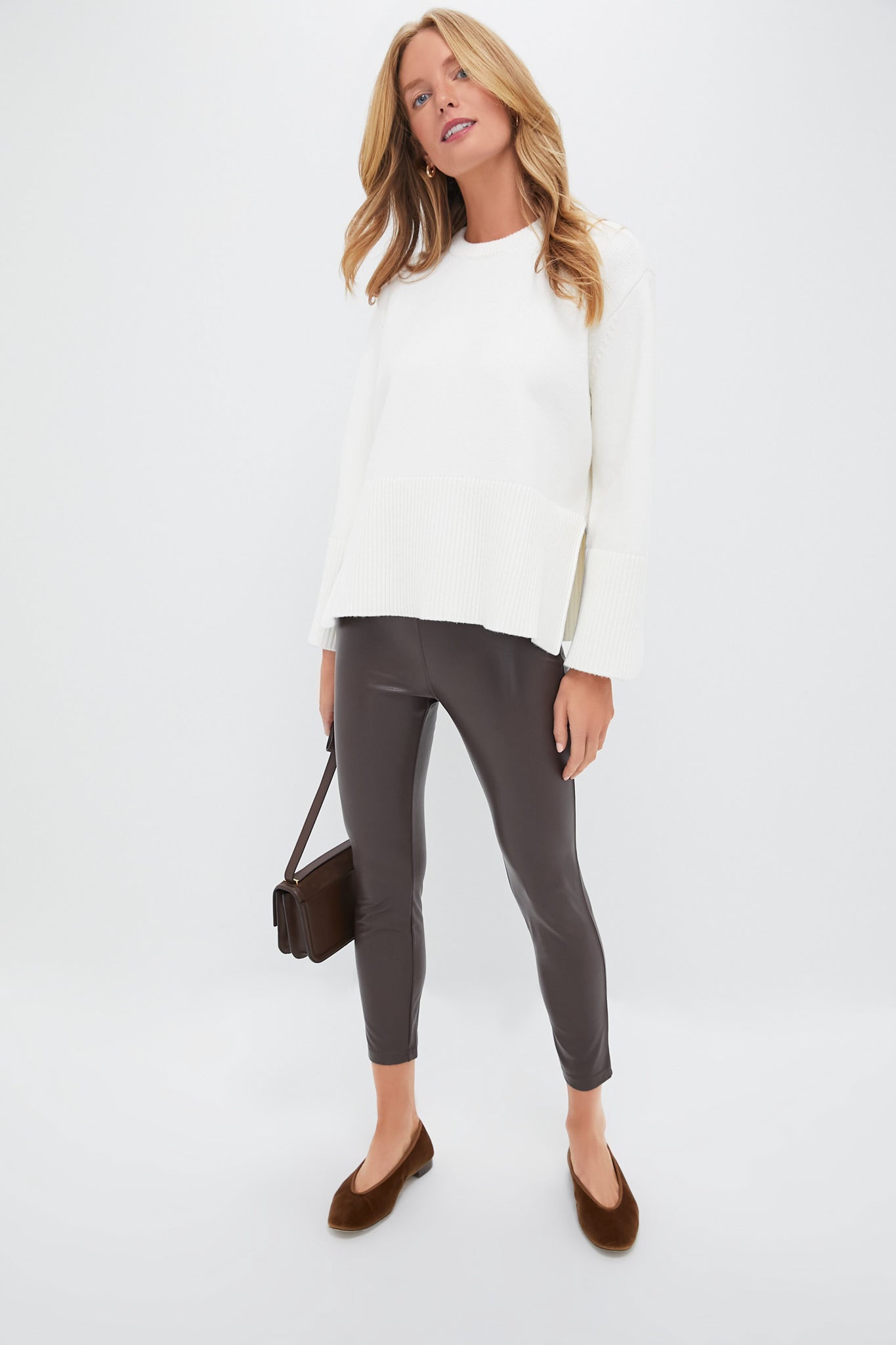 A woman stands against a plain background, wearing the Pomander Place Winter White Carington Sweater paired with dark brown leather-look leggings, brown flats, and a small brown shoulder bag for a classic silhouette.