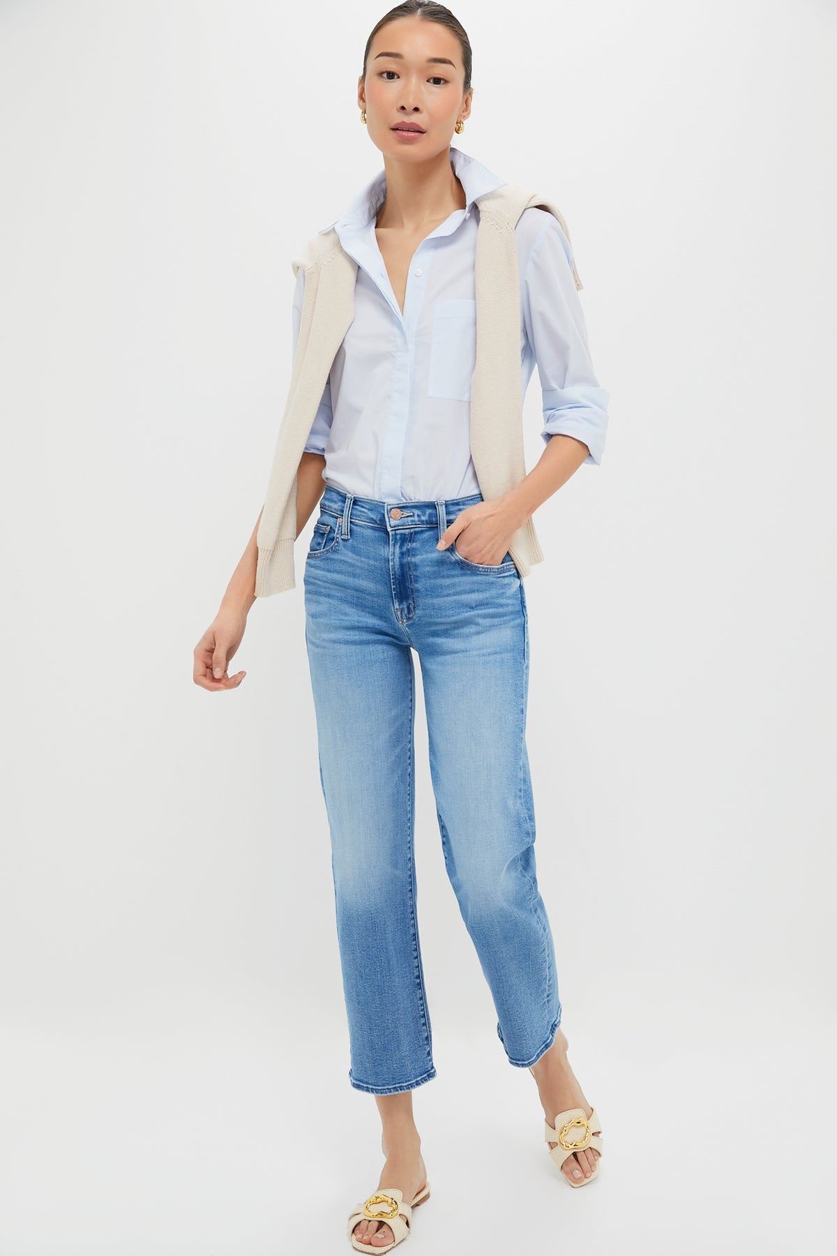 A person stands against a plain background wearing a light blue button-up shirt, a cream sweater over their shoulders, MOTHER mid-rise straight leg jeans, and beige sandals.