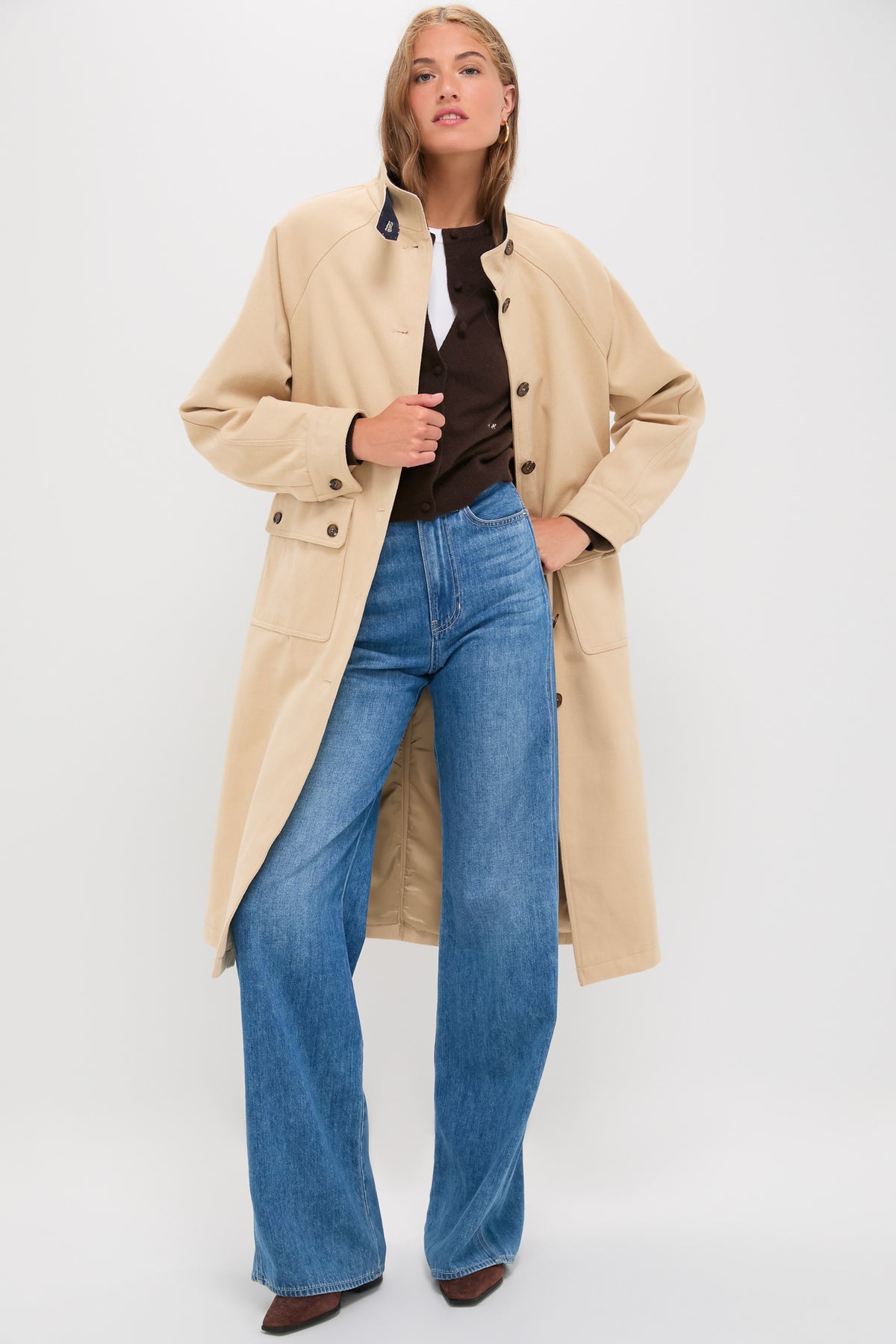 A woman stands wearing a long beige Line & Dot coat in brushed cotton twill over a dark top and blue wide-leg jeans with brown shoes.