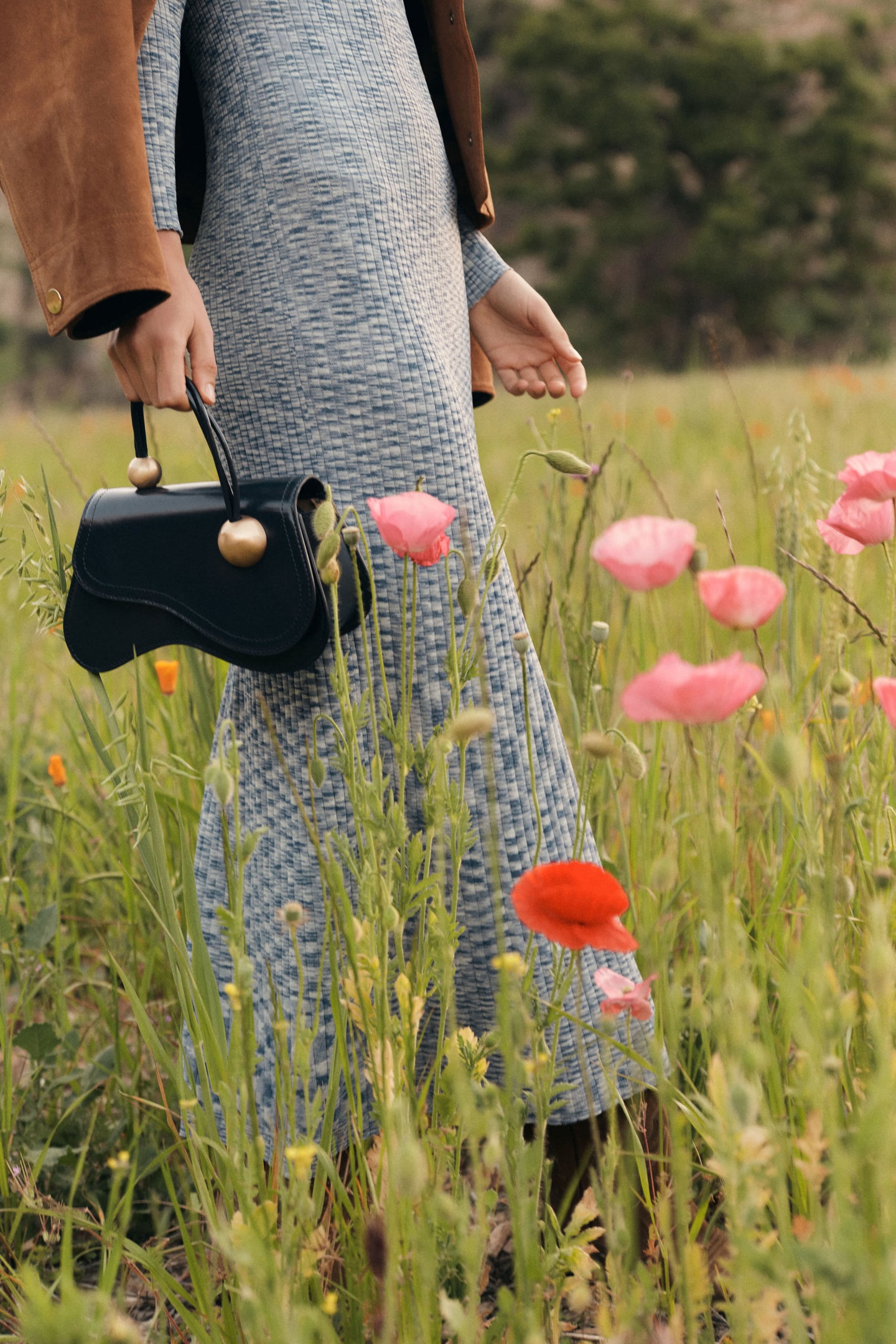 Woman carrying a small black handbag with circular gold hardware through a field of flowers. She's wearing a long blue knit dress with a brown jacket. 
