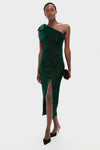 A person models a Saylor one-shoulder dark green shimmery dress with a thigh-high slit, paired with black heels and a black clutch, standing against a plain white background.