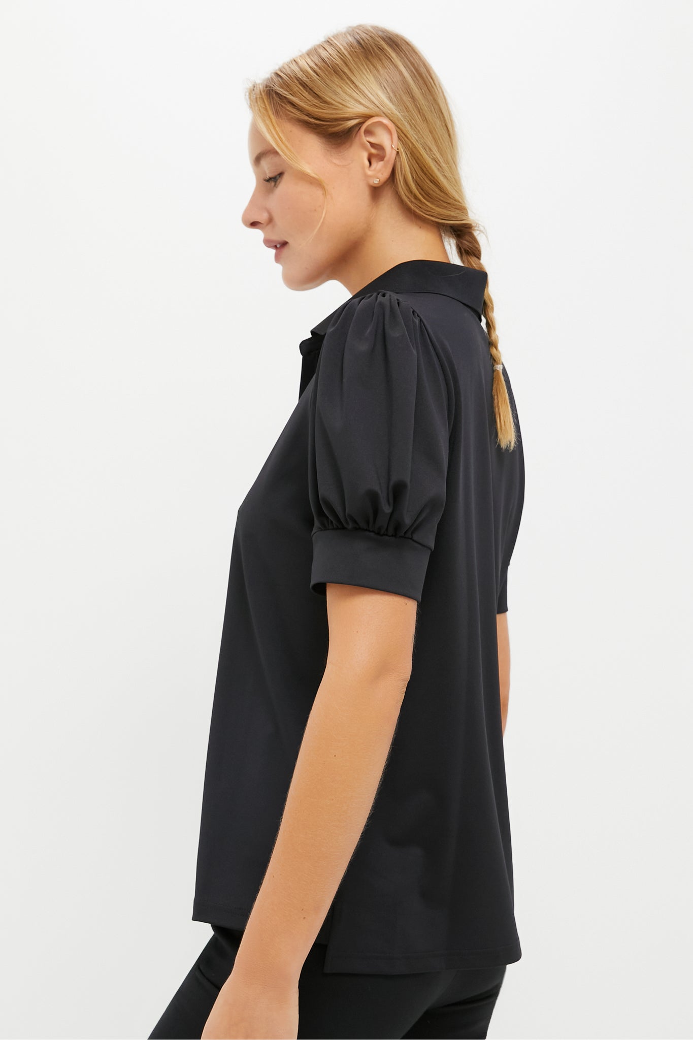 A woman stands in profile on a white background, wearing a black Tnuck Sport Puff Sleeve Caroline Polo with short puffed sleeves and collar. Hair in a simple braid. This stylish women’s golf polo features SPF protection for sunny days.