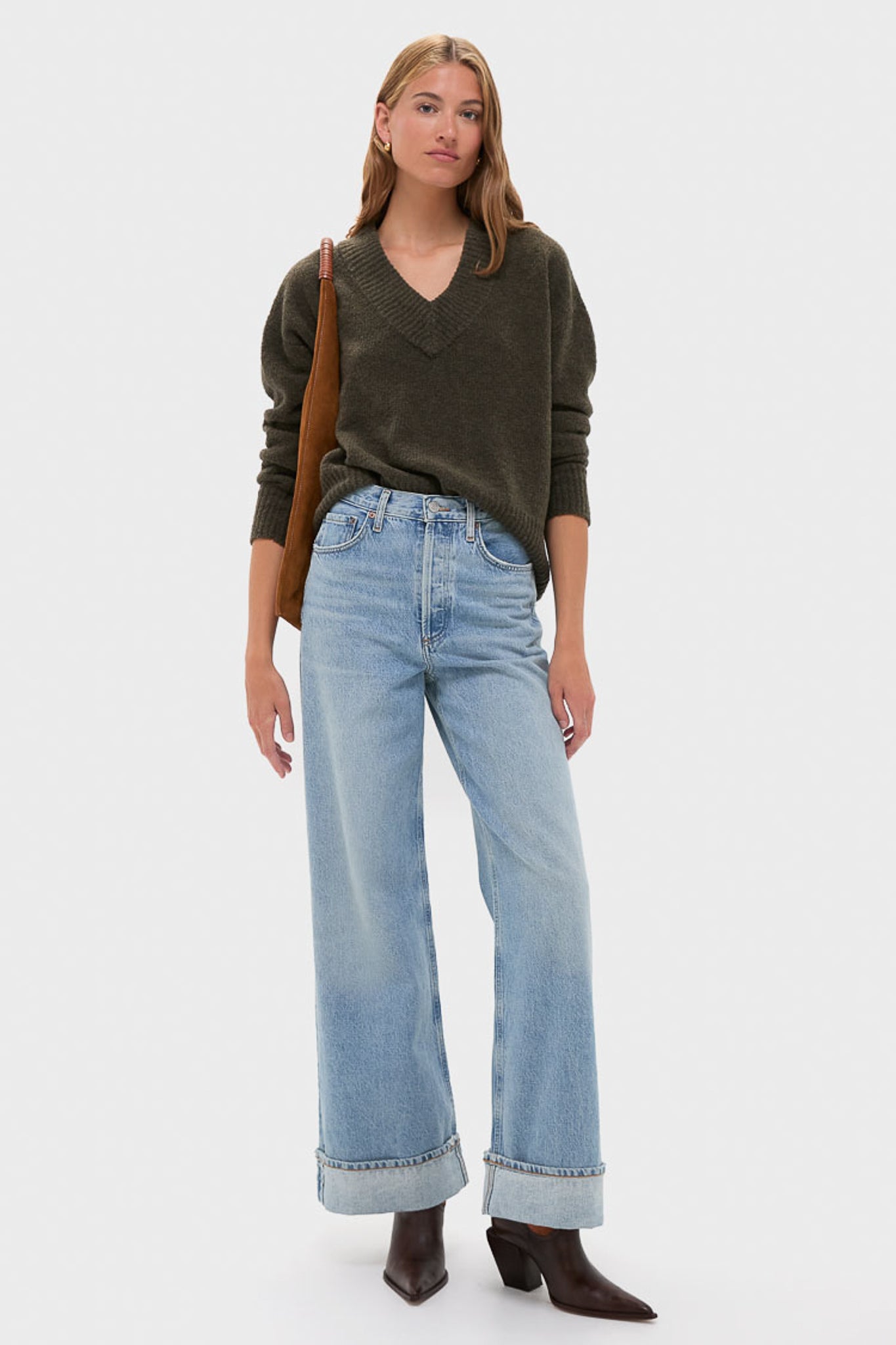 An individual styled in Emerson Fry’s oversized V-neck pullover, wide-leg cuffed jeans, brown heeled boots, and a brown shoulder bag stands against a plain light background.