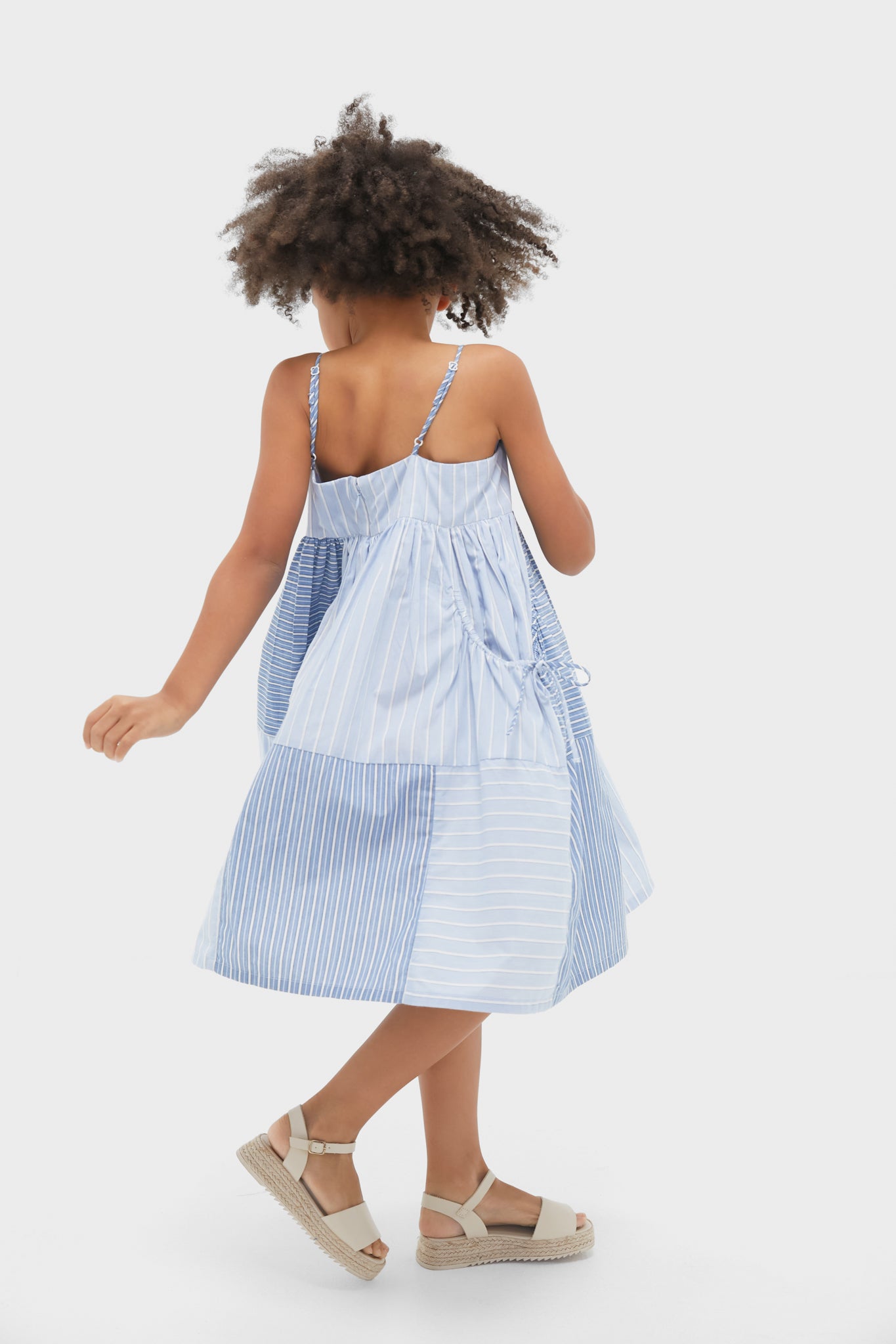 A young girl with curly hair wears a Sea New York Blue Helmi Stripe Tank Dress with oversized side pockets and beige sandals, standing against a plain white background.
