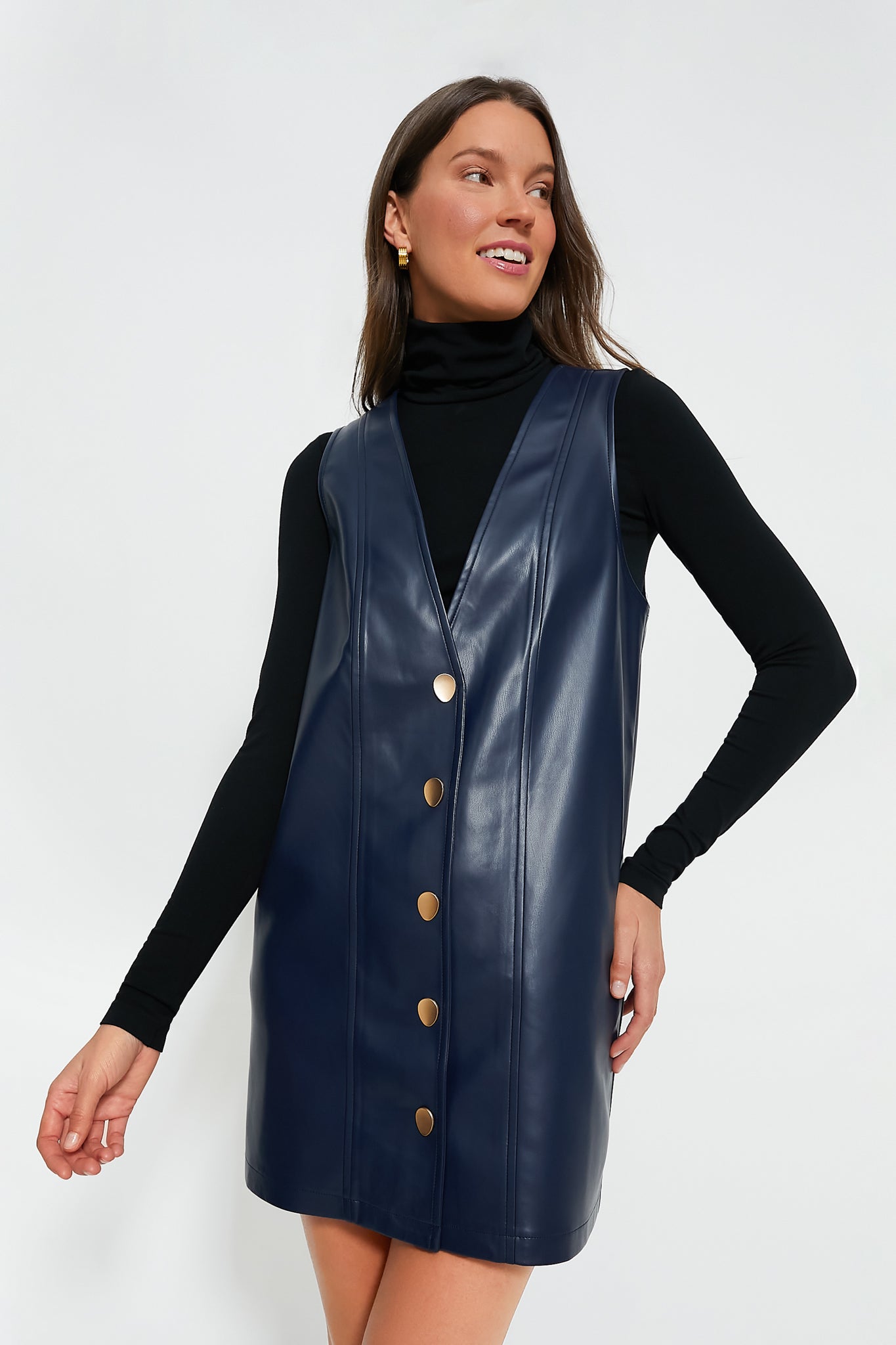 A woman models the Hyacinth House navy vegan leather pinafore dress with large gold buttons over a black turtleneck, highlighting its chic shift silhouette.