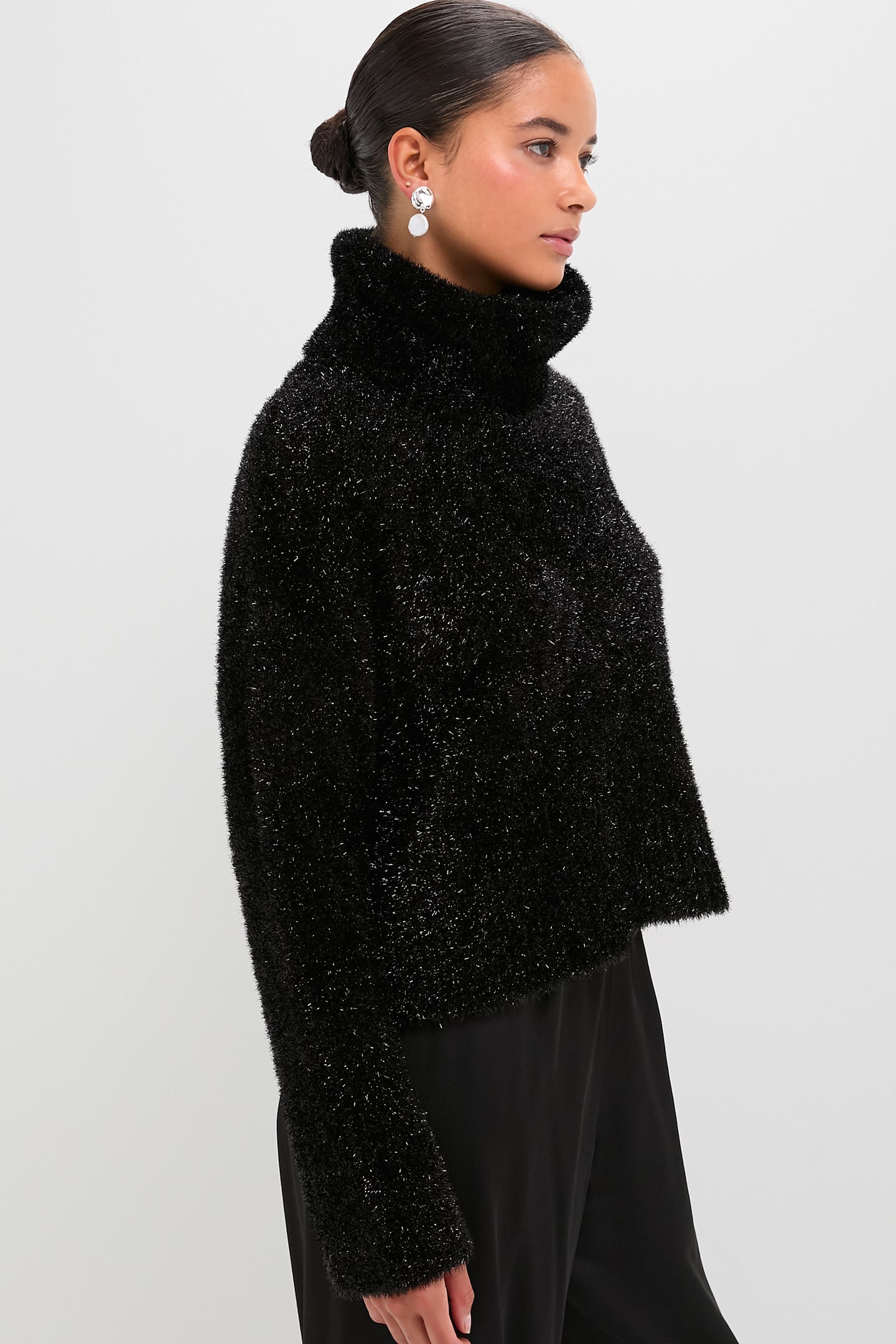 A woman wears a Hunter Bell black shimmering knit turtleneck sweater with black pants, standing against a plain white background.