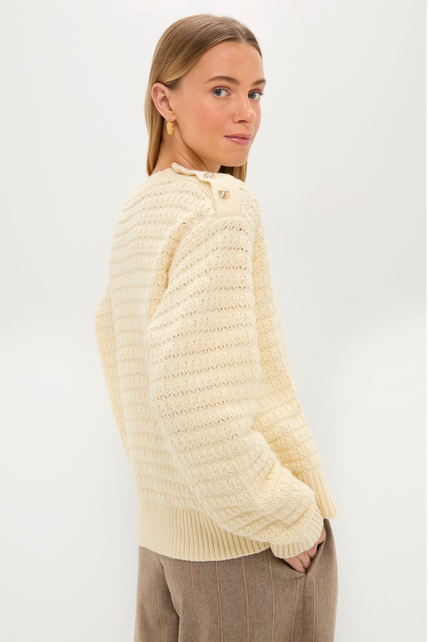A woman with straight, light brown hair wears a cream textured knit GANNI sweater and brown trousers, standing with hands in her pockets against a plain white background.
