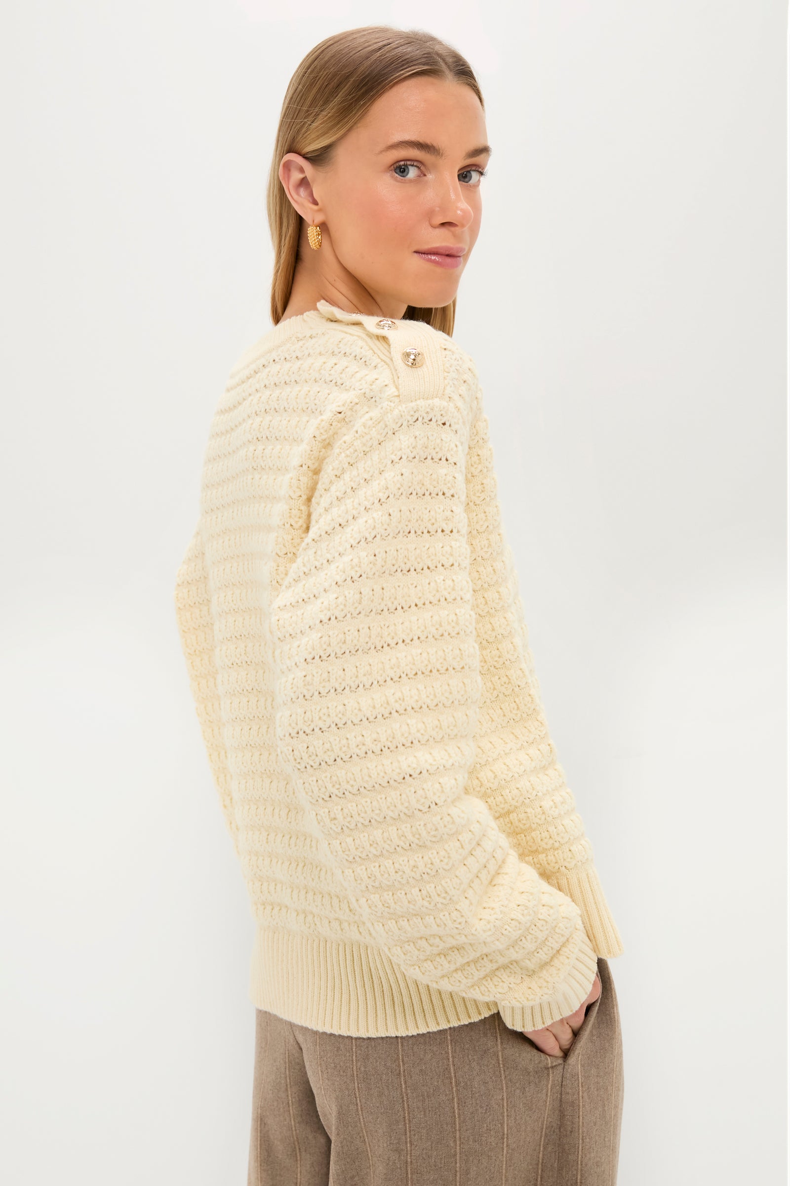 A woman with straight, light brown hair wears a cream textured knit GANNI sweater and brown trousers, standing with hands in her pockets against a plain white background.