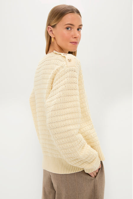 A woman with straight, light brown hair wears a cream textured knit GANNI sweater and brown trousers, standing with hands in her pockets against a plain white background.