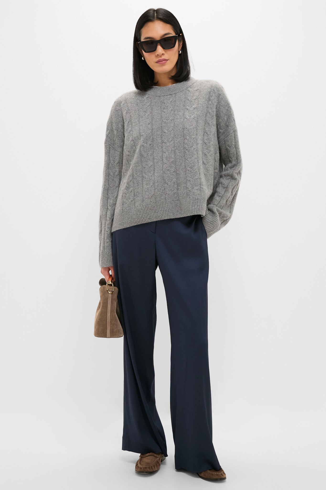 A woman in a gray cable-knit TWP sweater, navy viscose satin straight-leg pants, brown loafers, dark sunglasses, and a brown handbag stands against a plain light background.