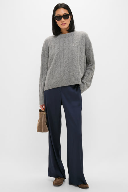 A woman in a gray cable-knit TWP sweater, navy viscose satin straight-leg pants, brown loafers, dark sunglasses, and a brown handbag stands against a plain light background.