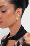 Woman wearing Gold Rici Earrings with gold-tone flower and pearl drop, paired with a floral top; a classic, effortless high-end accessory for any occasion.