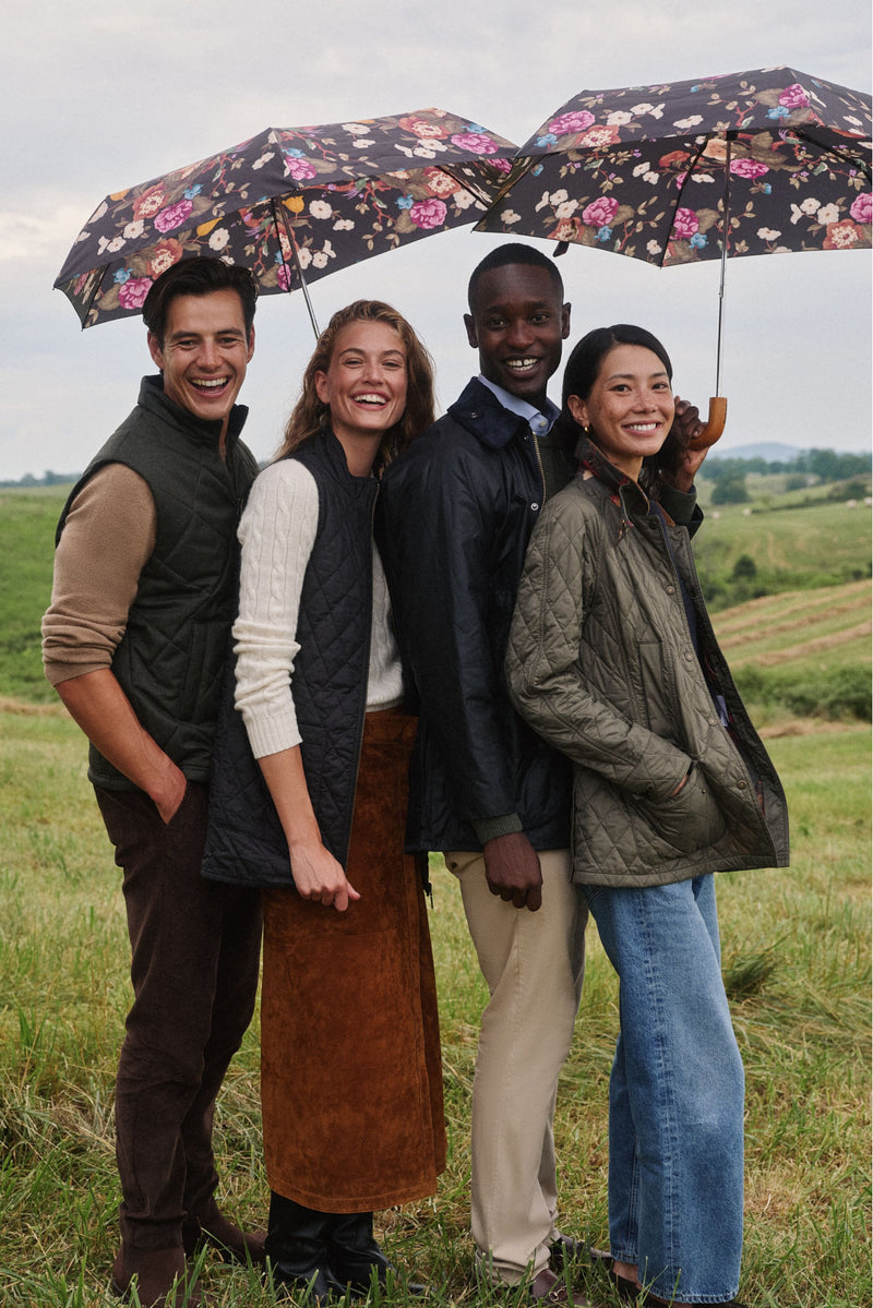 Four people stand outdoors in grassy fields, smiling under two Barbour x Tuckernuck umbrellas and wearing autumn jackets.