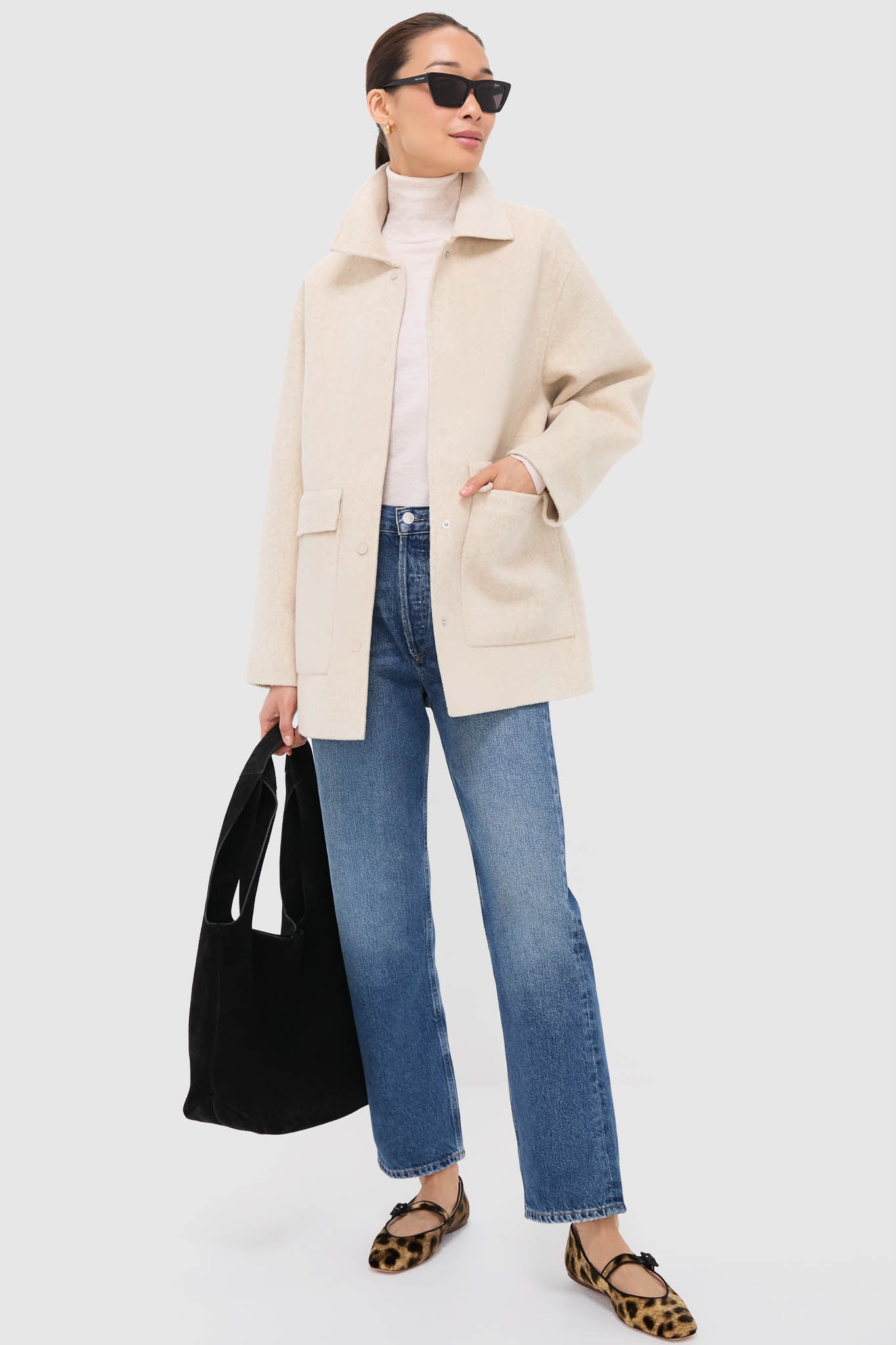 A woman in an AGOLDE cream jacket, white turtleneck, mid-rise straight leg jeans, and animal print flats holds a black tote bag and wears sunglasses against a plain background.