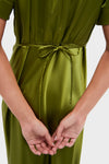 A person in a green vintage-inspired Tuckernuck dress is shown from the back, hands clasped and a thin belt tied in a bow at the waist.