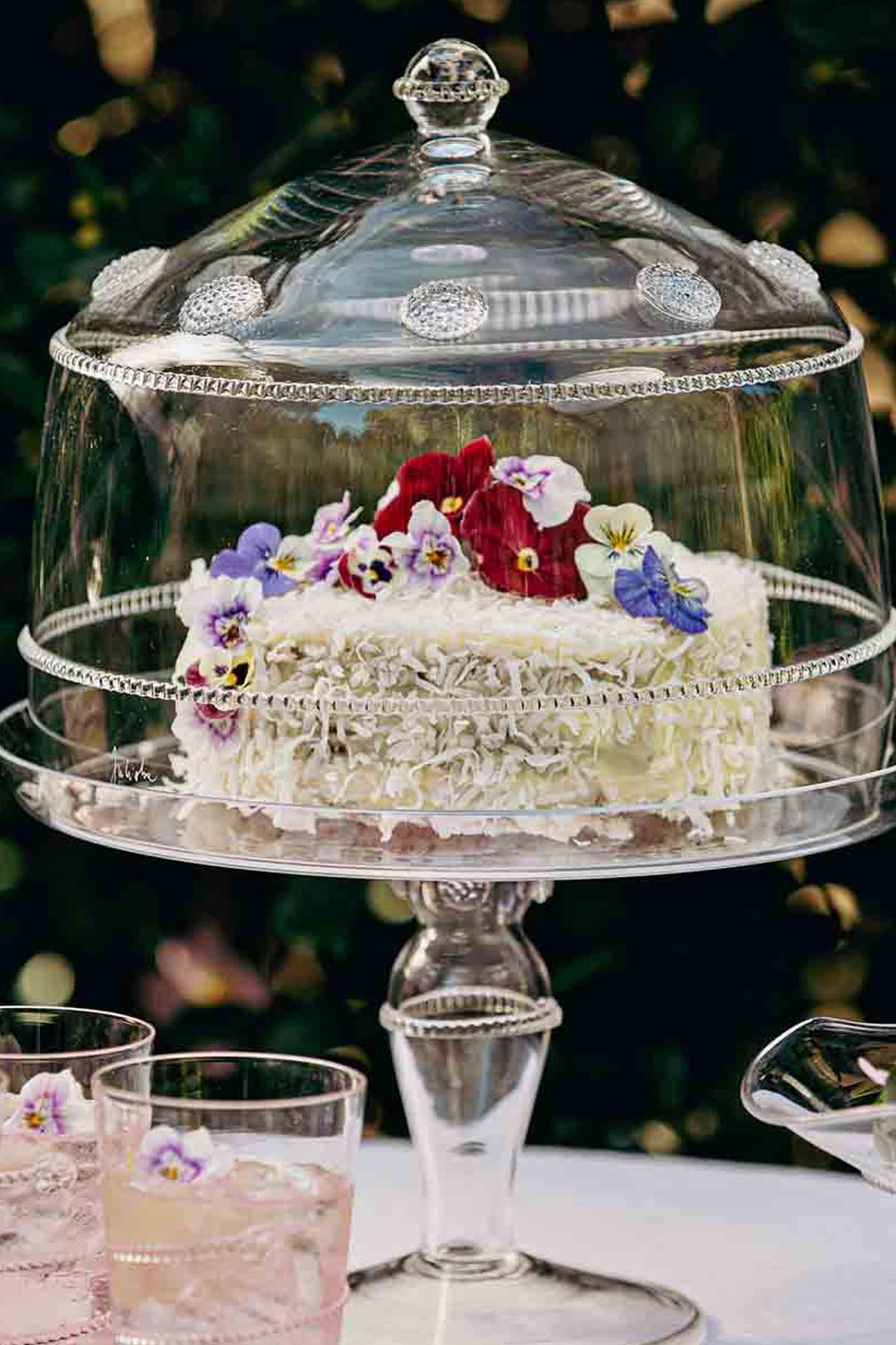 Isabella Cake Stand elegantly displays a coconut-frosted cake under a glass cloche, embodying classic, effortless high-end style for a fun, garden-style gathering.