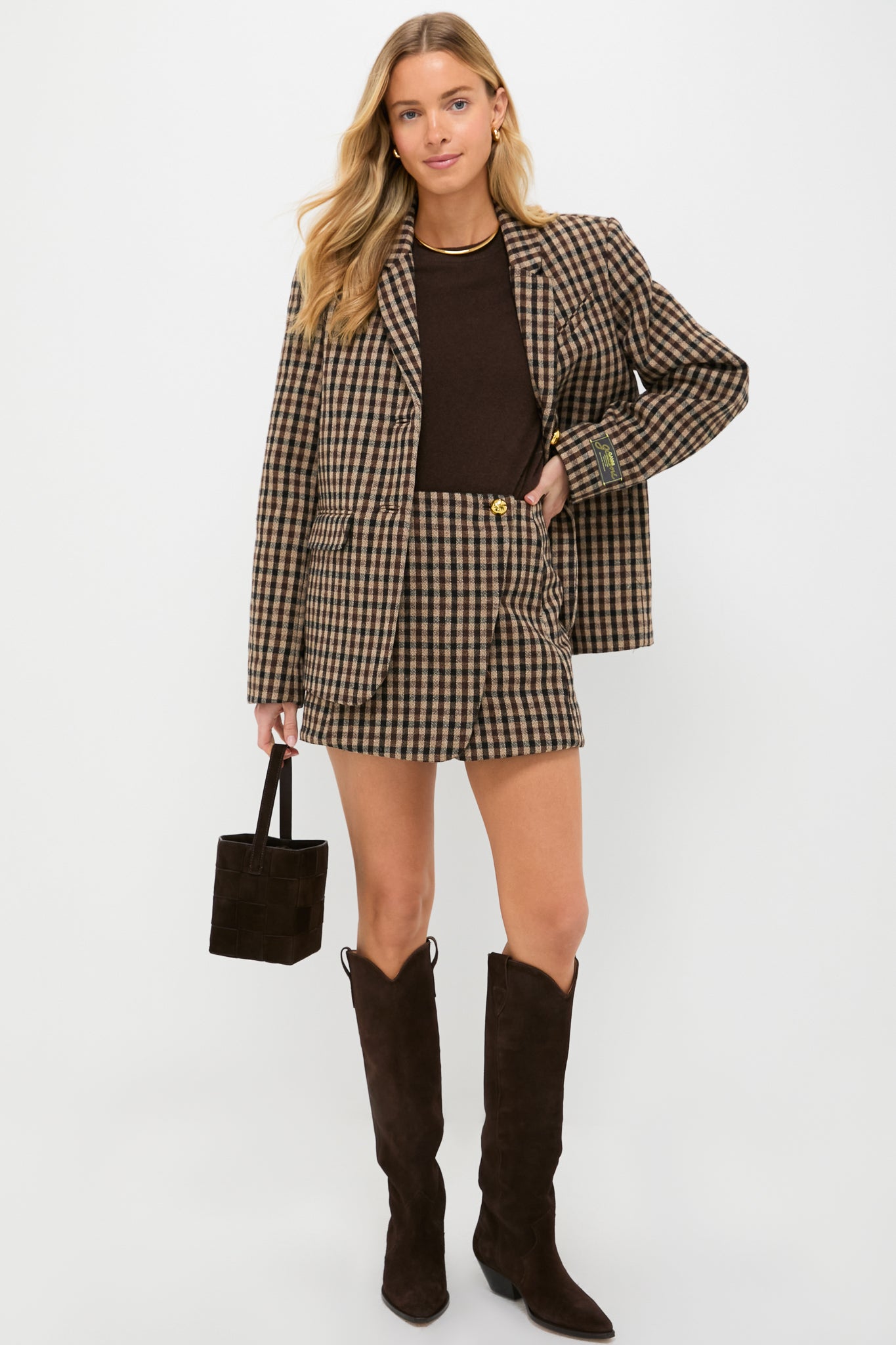 A woman stands in Scandinavian style, wearing a brown plaid blazer and matching shorts from GANNI, paired with a dark brown top, knee-high boots, and carrying a small brown handbag.