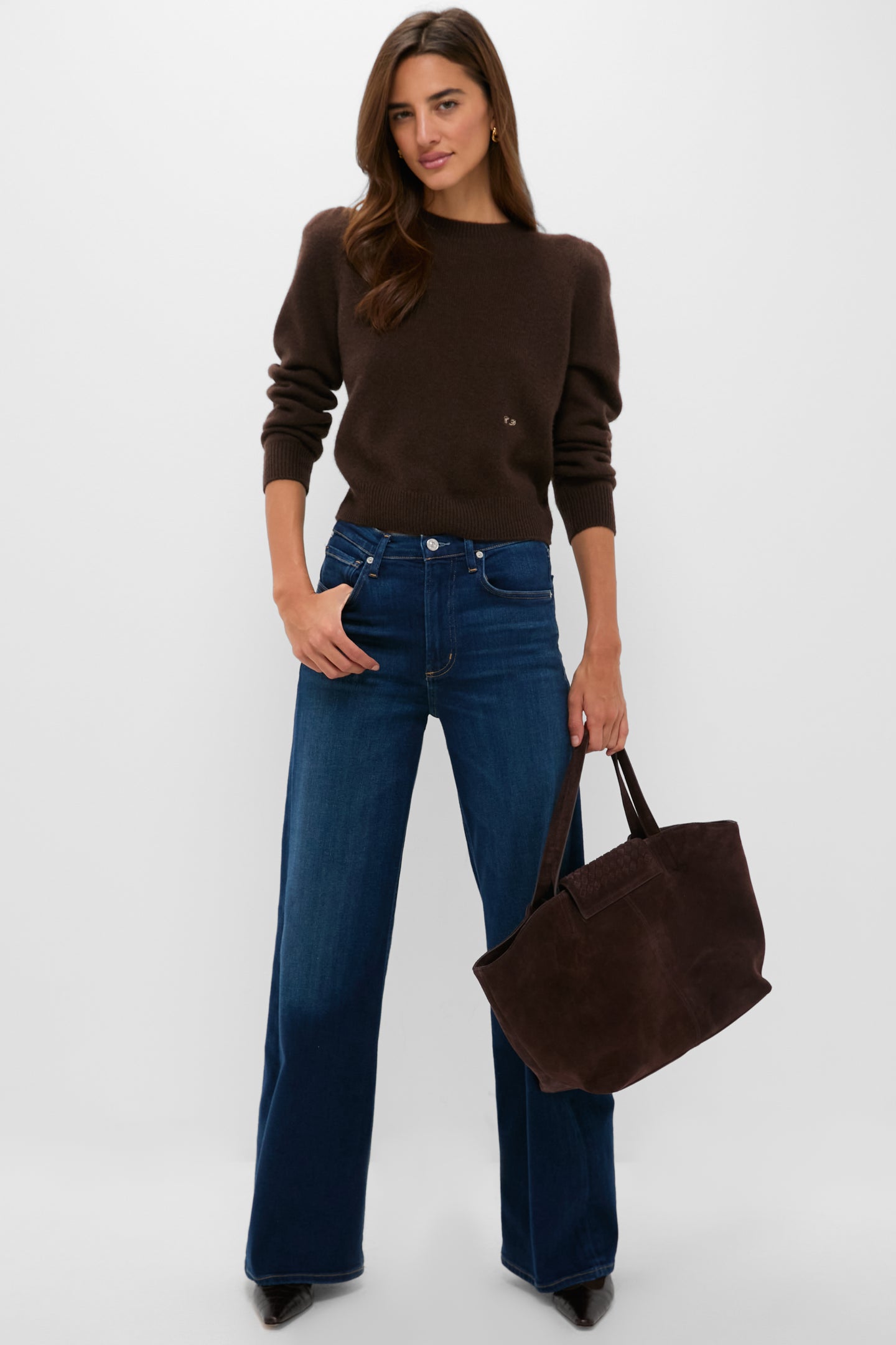 A woman in a Tuckernuck brown puff sleeve sweater and blue wide-leg jeans holds a dark brown tote bag, standing against a plain white background.