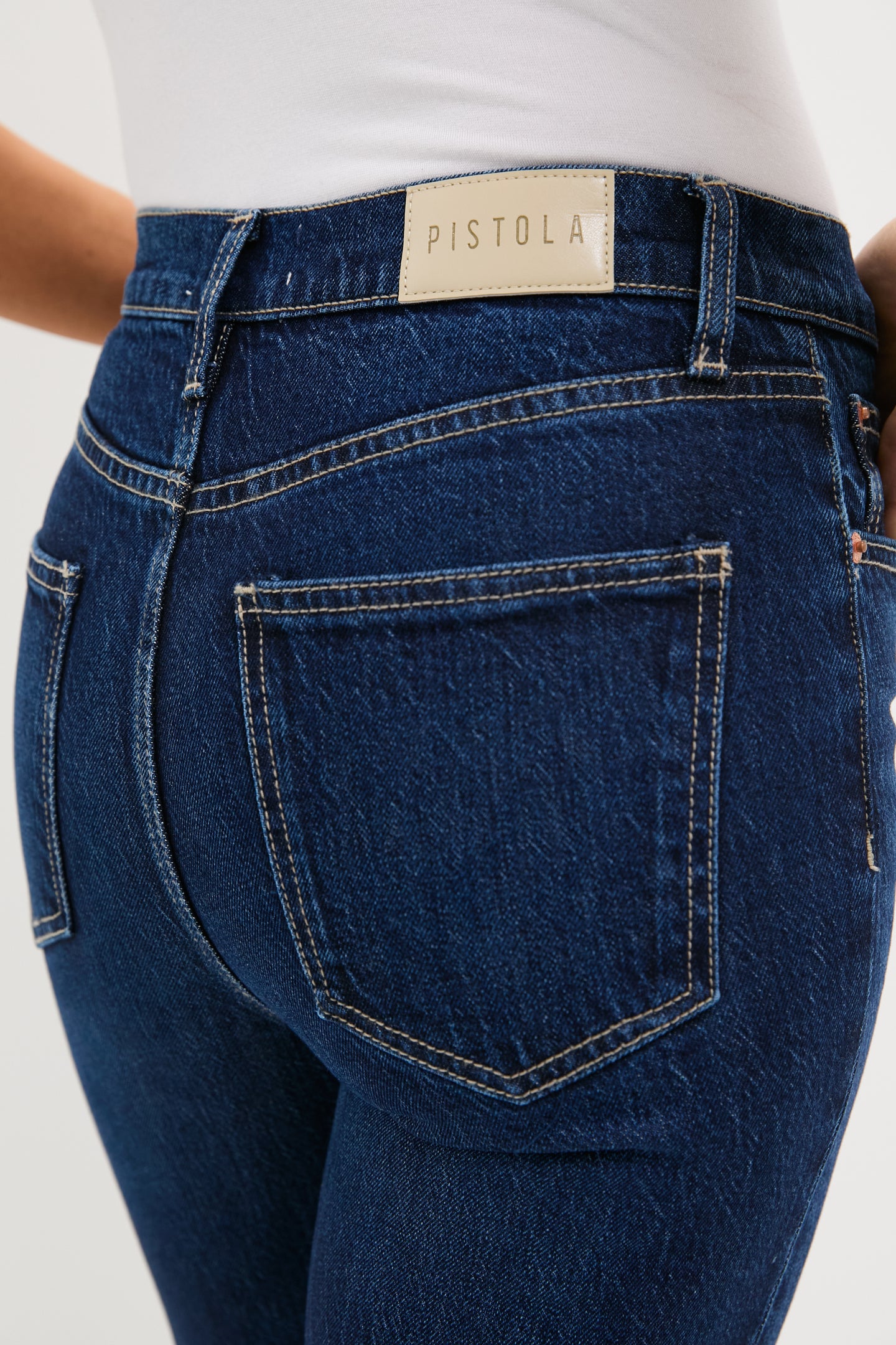 Close-up of a person wearing dark wash high rise slim straight jeans by Pistola Denim and a white top, highlighting the waistband, back pockets, and brand label.