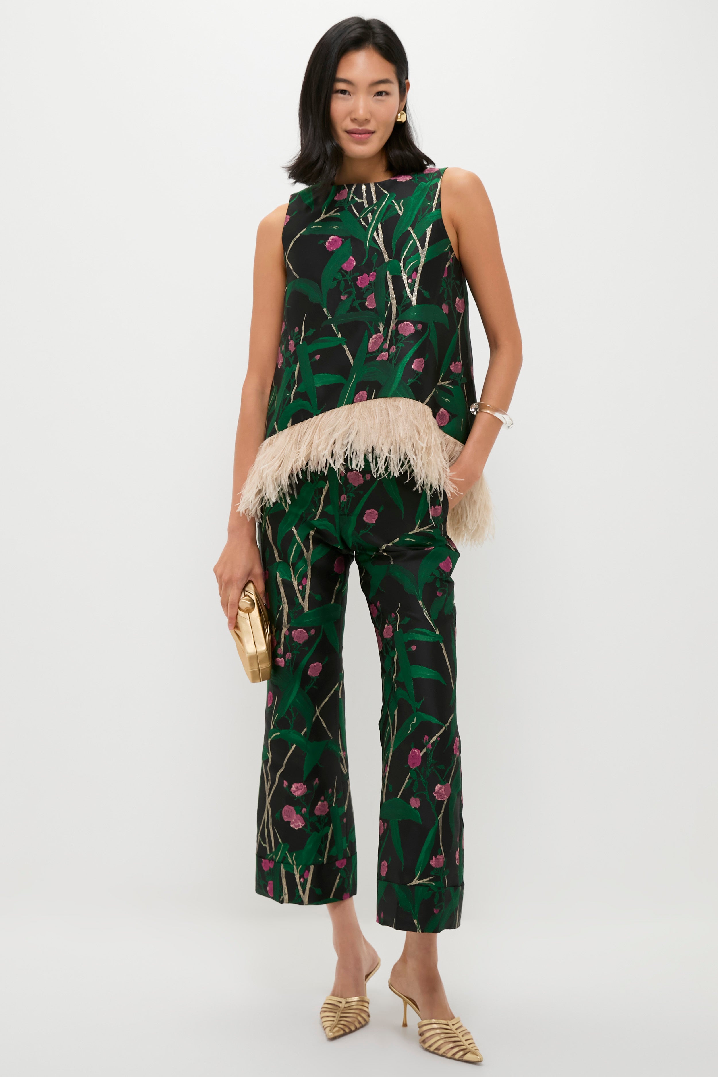 A woman wears a sleeveless, dark green La DoubleJ top with an ostrich feather-trimmed hem, matching pants, gold heels, and holds a small gold clutch against a plain white background.