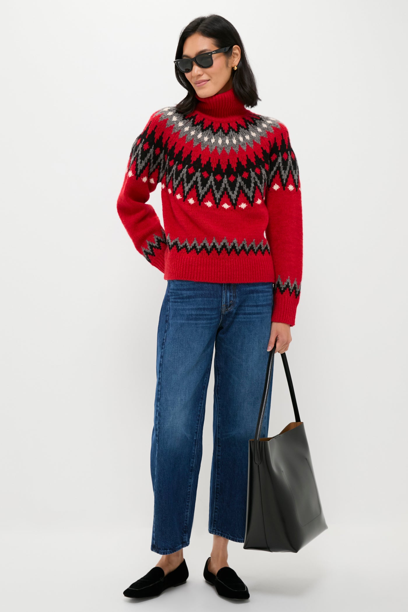 A woman wears a red Fair Isle sweater by Polo Ralph Lauren, blue jeans, black loafers, and sunglasses while holding a large black tote bag, standing against a plain white background.
